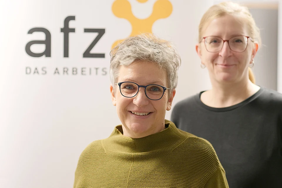 Zwei Frauen aus dem AFZ Team vor einem Plakat nit gelben AFZ Schriftzug.